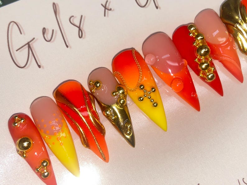May include: A set of long, pointed artificial nails with a gradient of colors, including yellow, orange, and red. Some nails feature gold embellishments, including beads and a cross pendant. The nails are arranged in a row.