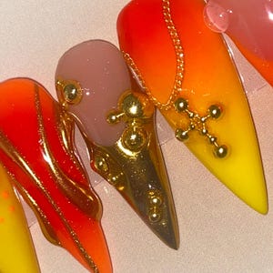 May include: Close-up of a set of long, pointed acrylic nails. The nails feature a gradient of red, orange, and yellow, with gold accents. One nail has a gold chain and cross charm. Other nails have gold embellishments.