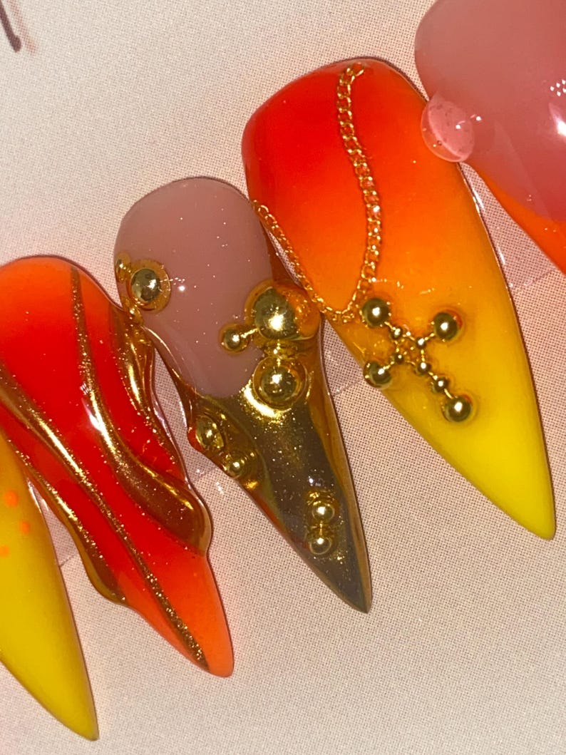 May include: Close-up of a set of long, pointed acrylic nails. The nails feature a gradient of red, orange, and yellow, with gold accents. One nail has a gold chain and cross charm. Other nails have gold embellishments.