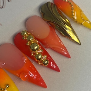 May include: A collection of stiletto-shaped artificial nails in vibrant colors. The nails feature a gradient of yellow, orange, and red hues, with gold embellishments. Some nails have gold accents, while others have a gold metallic design.
