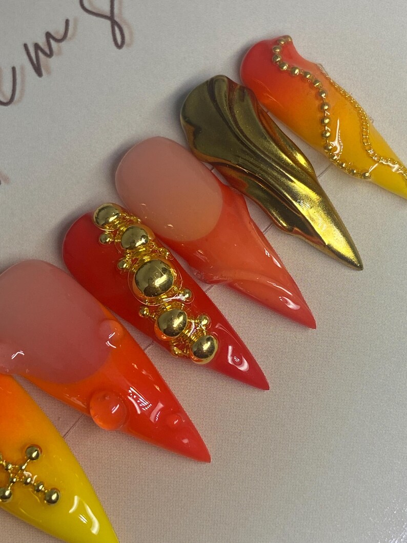 May include: A collection of stiletto-shaped artificial nails in vibrant colors. The nails feature a gradient of yellow, orange, and red hues, with gold embellishments. Some nails have gold accents, while others have a gold metallic design.