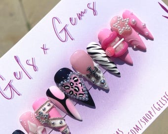 May include: A set of long, pointed artificial nails in various designs. The nails feature pink, black, and white colors, with embellishments like pearls, rhinestones, and animal prints. The text "Gels x Gems" is visible.