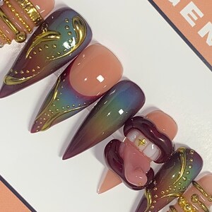 May include: A set of long, pointed artificial nails in various designs. Some nails feature a gradient of colors, while others have gold embellishments and a 3D design of a mouth with teeth. The nails are displayed on a white surface.