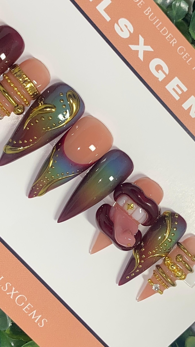 May include: A set of long, pointed artificial nails in various designs. Some nails feature a gradient of colors, while others have gold embellishments and a 3D design of a mouth with teeth. The nails are displayed on a white surface.