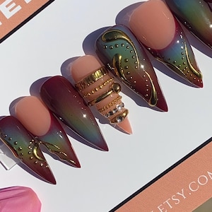 May include: Stiletto-shaped false nails with a gradient of colors, including red, green, and gold. One nail features multiple gold rings with rhinestones. The nails are arranged on a white surface with a pink and peach-colored card.
