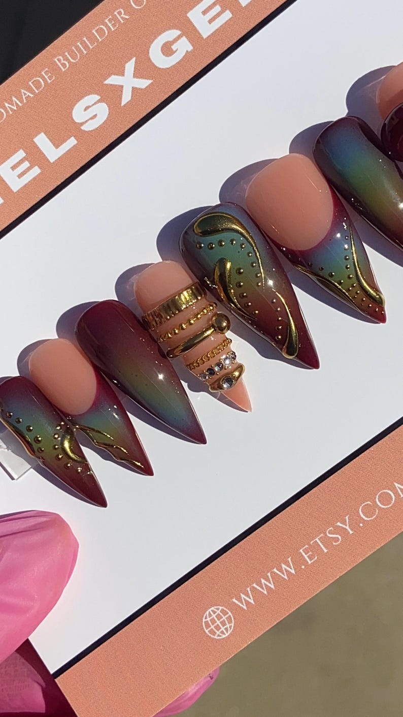 May include: Stiletto-shaped false nails with a gradient of colors, including red, green, and gold. One nail features multiple gold rings with rhinestones. The nails are arranged on a white surface with a pink and peach-colored card.
