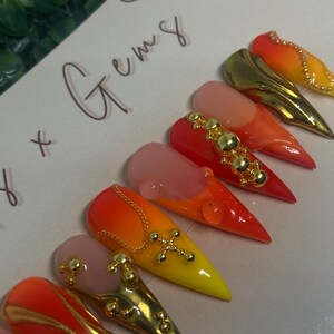 May include: A collection of stiletto-shaped artificial nails with a gradient of colors, including orange, yellow, and pink. Some nails feature gold embellishments like chains, studs, and crosses. The text "x Gems" is handwritten on the background.