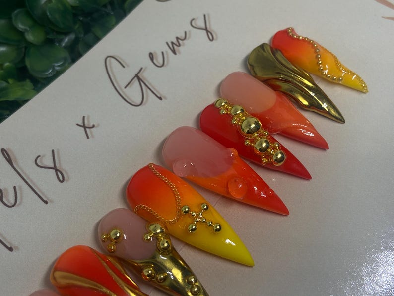 May include: A collection of stiletto-shaped artificial nails with a gradient of colors, including orange, yellow, and pink. Some nails feature gold embellishments like chains, studs, and crosses. The text "x Gems" is handwritten on the background.