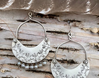 May include: A pair of silver hoop earrings with a crescent moon design and pearl accents. The earrings are handmade and feature a delicate floral pattern.