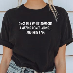 Puede incluir: Camiseta negra con el texto blanco: "ONCE IN A WHILE SOMEONE AMAZING COMES ALONG... AND HERE I AM." La camiseta la lleva una persona con pelo rubio y vaqueros azules. La camiseta tiene las mangas dobladas.