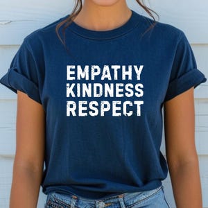 Puede incluir: Camiseta azul marino con las palabras "EMPATHY KINDNESS RESPECT" impresas en letras blancas. La camiseta tiene las mangas enrolladas y la lleva una persona. La persona lleva vaqueros azules.
