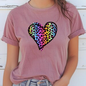 May include: A mauve-colored t-shirt featuring a large heart design. The heart is filled with a colorful leopard print pattern in shades of pink, yellow, blue, and green, outlined in black. The shirt has short sleeves and a relaxed fit.