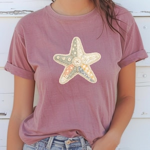 May include: A dusty rose-colored t-shirt featuring a starfish design. The starfish is made of various patterned fabrics and adorned with buttons. The shirt has short sleeves and a relaxed fit, perfect for casual wear.