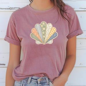 May include: A dusty rose-colored t-shirt featuring a seashell design. The seashell is composed of various fabric patterns, including stripes, florals, and gingham, in shades of orange, green, blue, and yellow. The shirt has short sleeves and a relaxed fit.