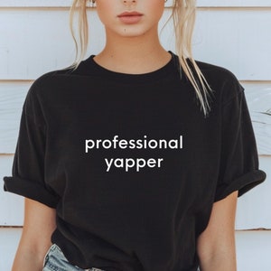 Può includere: Una maglietta nera con la scritta "professional yapper" stampata in bianco sul davanti.