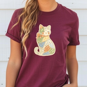 Puede incluir: Camiseta burdeos con un diseño de gato patchwork. El gato tiene un lazo y está hecho de varios parches de tela coloridos. La camiseta tiene mangas cortas y un corte relajado.
