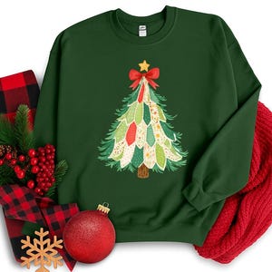 May include: A dark green sweatshirt with a Christmas tree design, a red bow, and a star. The tree is decorated with colourful shapes. A red scarf, a red and black plaid blanket, and a red bauble are also present.