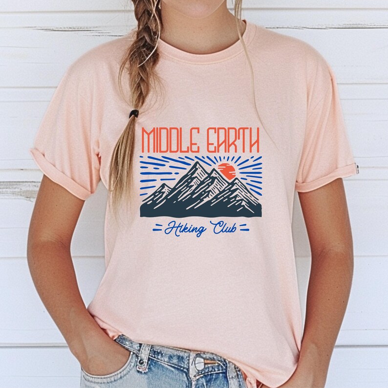 Op de afbeelding: Een roze T-shirt met een grafisch ontwerp van bergen, een zon en de tekst "Middle Earth Hiking Club".
