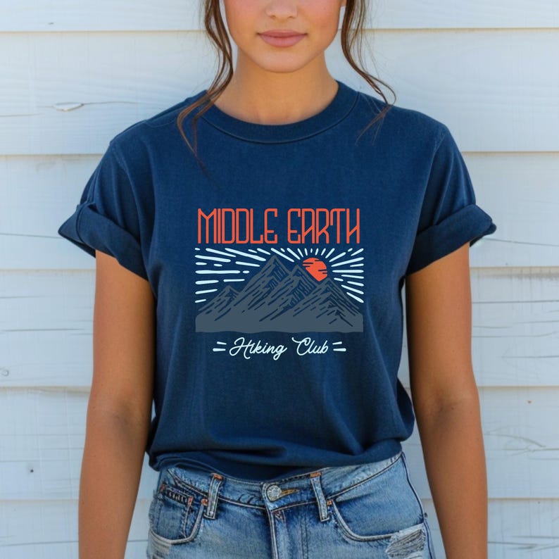 Op de afbeelding: Een marineblauw t-shirt met een afbeelding van bergen en een zon met de tekst "Middle Earth Hiking Club".
