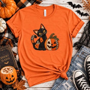 May include: An orange short-sleeved t-shirt featuring a graphic of a black cat with patchwork patterns sitting next to a jack-o'-lantern pumpkin. The cat has yellow eyes and the pumpkin has a black and white checkered pattern on its side.