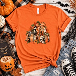 Op de afbeelding: Een oranje t-shirt met korte mouwen en een grafische print van drie pompoenen met diverse patronen zoals ruiten, sterren en manen. De pompoenen zijn versierd met strikken, bladeren en kleine labels met de tekst Trick or Treat en Boo.