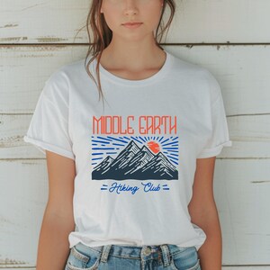 Op de afbeelding: Wit T-shirt met een afbeelding van bergen en een zon met de tekst "Middle Earth - Hiking Club -" erop gedrukt.