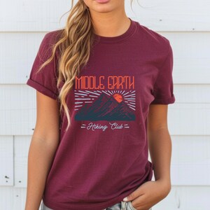 Op de afbeelding: Bordeauxkleurig T-shirt met een afbeelding van bergen en een zon met de tekst "Middle Earth Hiking Club" gedrukt op de voorkant.