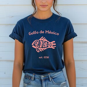 May include: A navy blue t-shirt with a pink fish graphic and the text "Golfo de M&eacute;xico est. 1550".