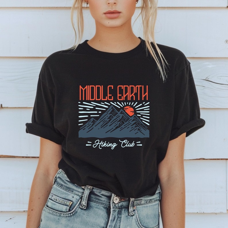 Op de afbeelding: Zwart T-shirt met een grafisch ontwerp van bergen, een zon en de tekst "Middle Earth Hiking Club".