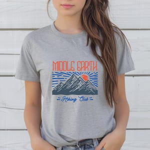 Op de afbeelding: Grijs T-shirt met een blauwe en oranje afbeelding van bergen en een zon met de tekst "Middle Earth Hiking Club".