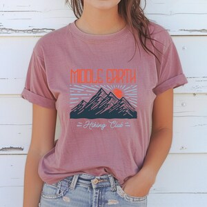 Op de afbeelding: Een mauve t-shirt met een afbeelding van bergen en een zon met de tekst "Middle Earth Hiking Club".