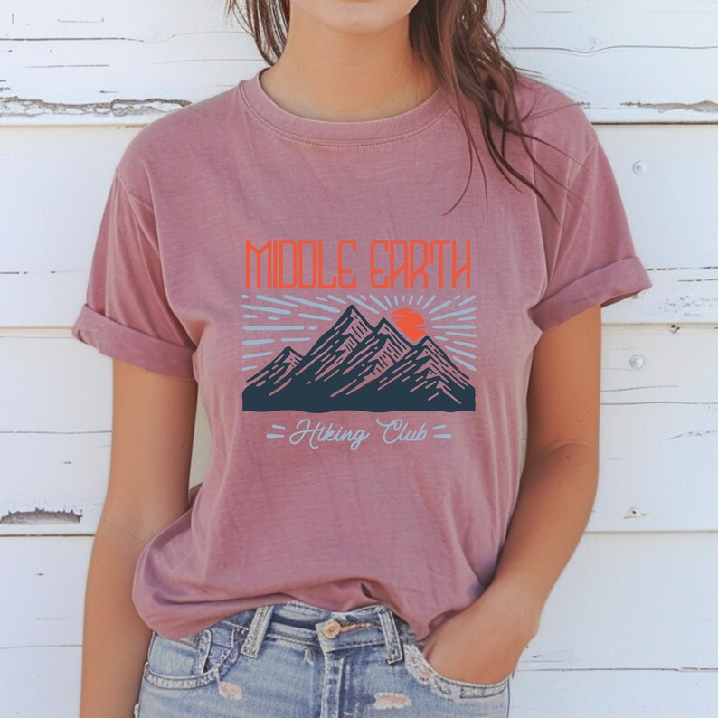 Op de afbeelding: Een mauve t-shirt met een afbeelding van bergen en een zon met de tekst "Middle Earth Hiking Club".