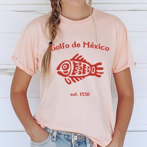 May include: A pink t-shirt with a red stylized fish and the text "Golfo de M&eacute;xico est. 1550".