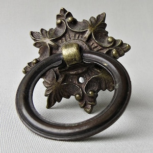 May include: Antique bronze drawer pull with a decorative floral backplate and a circular ring handle. The hardware has a vintage aesthetic, with a dark bronze finish and brass accents. The pull is designed for furniture and cabinets.