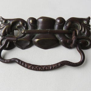 May include: Antique bronze drawer pull with ornate, stylized floral and foliate designs. The pull features a curved handle and decorative elements on either side. The hardware is likely made of cast metal and has a dark, aged patina.