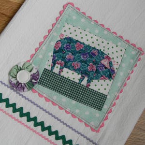 May include: White kitchen towel with a patchwork pig design. The pig is made of floral fabric and set on a green and white polka dot background. The towel has a pink scalloped edge and decorative green and purple trim.
