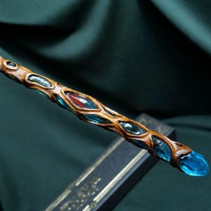 Puede incluir: Una varita de madera con un mango marrón texturizado y nudoso, que presenta una serie de inserciones translúcidas de color azul claro similares a cristales a lo largo de su cuerpo, con una punta de cristal azul y un acento rojo.