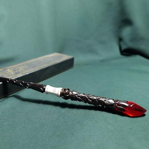 May include: A dark brown wand featuring a textured handle with a skull motif, a white band with a small symbol, and a translucent red crystal tip. The wand is positioned next to a long, narrow, dark rectangular box with gold-coloured trim.
