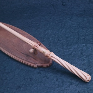 Puede incluir: Una varita de madera de color claro con un mango en espiral y una talla en forma de estrella cerca del centro, apoyada sobre un soporte de exhibición ovalado de madera marrón oscuro con dos pequeñas clavijas de soporte.