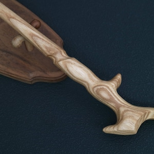 May include: A light-colored wooden wand with a carved handle and a curved end, resting on a wooden display stand. The wand has a natural wood grain pattern and is angled diagonally across the frame.