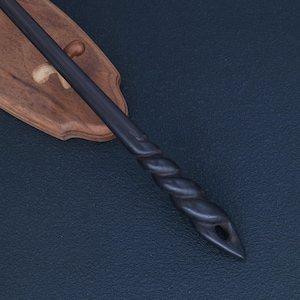 May include: A dark, carved wooden wand with a twisted handle and a hole at the tip, resting on a wooden display stand. The wand is dark gray, and the stand is a light brown color. The background is a dark blue surface.