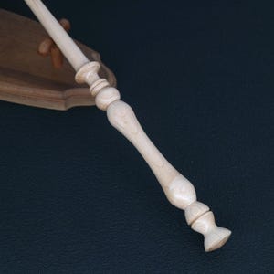 May include: A light-colored wooden wand with a detailed handle and a flared base. The wand is angled diagonally against a dark background, showcasing its carved design and smooth finish. A wooden stand is visible in the background.