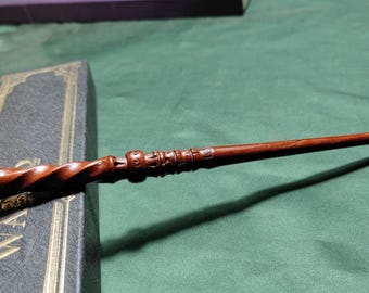 Może przedstawiać: Ciemnobrązowa drewniana różdżka z misternymi rzeźbieniami i skręconą rączką spoczywa na ciemnej książce ze złotymi literami. Projekt różdżki zawiera szczegółowe wzory i gładki, zwężający się koniec. Tło to jednolita zielona powierzchnia.