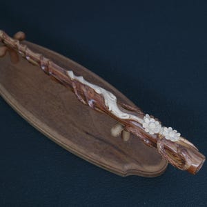 May include: A handcrafted wooden wand with intricate carvings, featuring a light-colored floral design and a dark brown finish. The wand rests on a wooden display stand, showcasing its detailed craftsmanship.