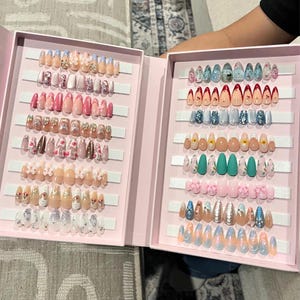May include: A pink box with rows of colorful fake nails. The nails are arranged in different designs and colors, including pink, blue, green, and white. The nails are decorated with glitter, flowers, bows, and other embellishments.