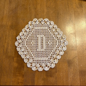 May include: Cream-colored, square doily with a scalloped edge. The doily is made of crocheted yarn, with an open, lacy design. The doily is approximately 8 inches square. The doily is placed on a wooden surface.