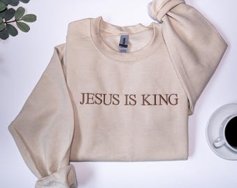 May include: A beige crewneck sweatshirt with the embroidered text "JESUS IS KING" in brown. The sweatshirt has long sleeves with rolled cuffs. A coffee cup and saucer are in the bottom right corner.
