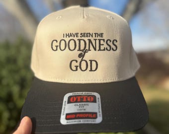 May include: A beige baseball cap with a black brim. The front of the cap has black embroidered text that reads "I have seen the goodness of God". The back of the cap has a black label with white text that reads "Premium Fabric Otto Classic Fit OSFM Mid Profile Maximum Comfort".