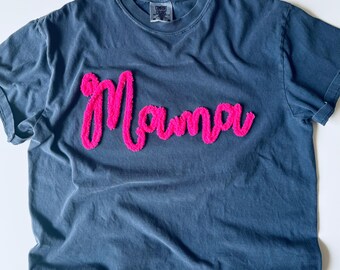 May include: A slate blue short-sleeved t-shirt featuring the word Mama in bright pink fuzzy chenille lettering across the chest. The shirt has a crew neckline and a Comfort Colors brand label visible on the inside of the collar area.