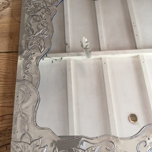 May include: Silver-toned rectangular mirror with an ornate, embossed border featuring a repeating leaf and vine design. The mirror's frame has a wavy edge and a reflective surface, suitable for home decor.